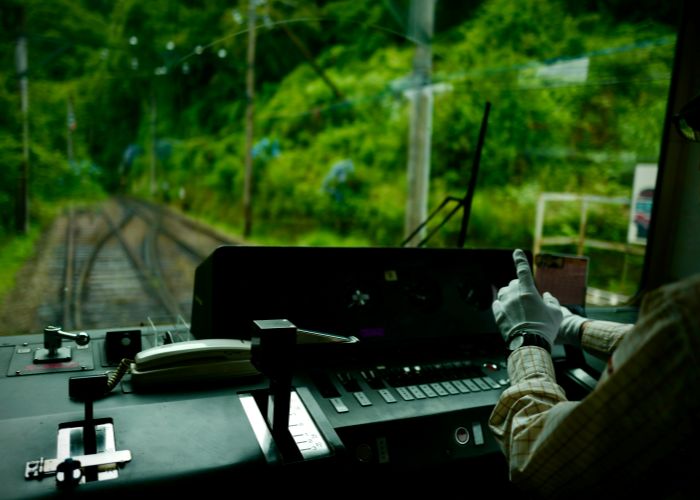 A Japanese train driver wearing white gloves and driving a train through an area of green trees.